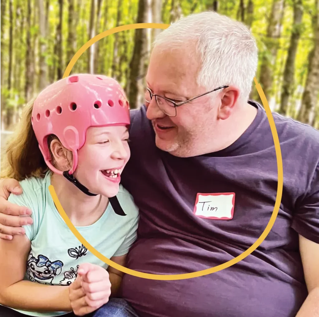 A man wearing glasses and a “Tim” name tag hugs a smiling girl in a pink helmet. They are sitting together outdoors with trees in the background, celebrating belonging and connection among children with disabilities.